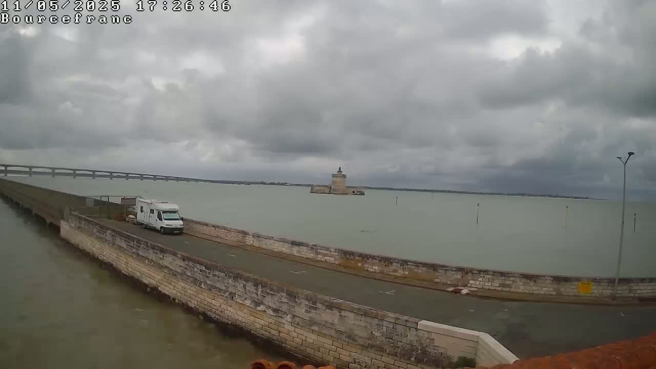 A white camper van is parked on a stone roadway next to a body of water, under an overcast sky, with a distant stone tower and bridge visible.