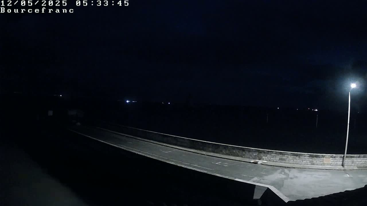 A dark, nighttime scene shows a paved roadway on a low wall overlooking a dark body of water under a clear, starless sky, lit by a single lamppost.