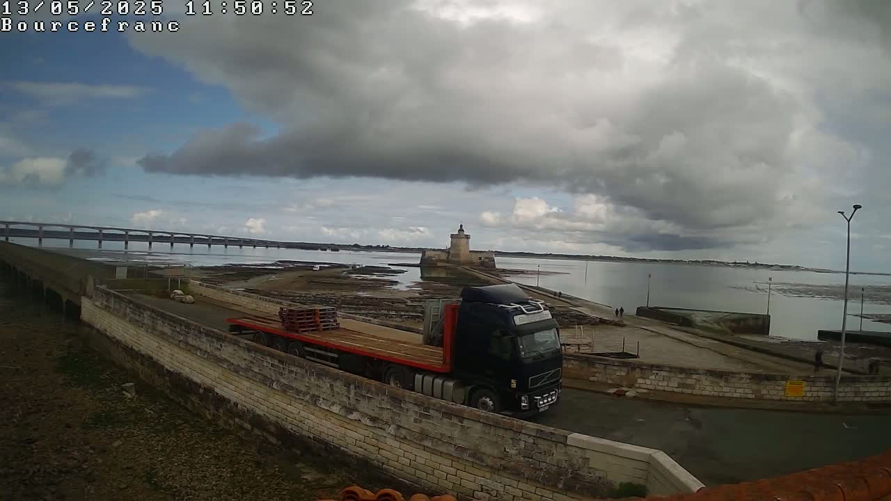 A dark-colored semi-truck carrying pallets is parked near a stone wall overlooking a body of water under a cloudy sky, with a distant bridge and a stone tower visible.