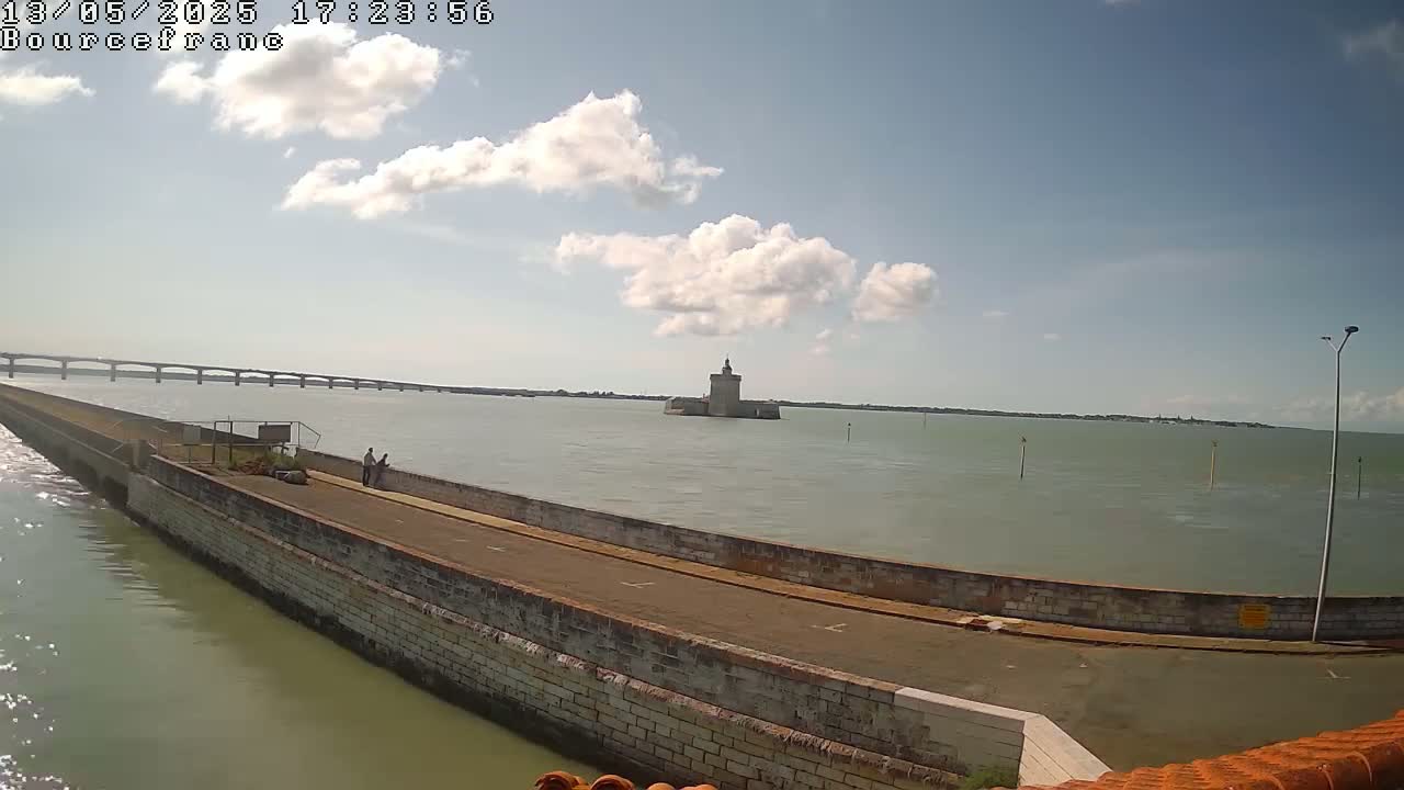 A stone seawall extends into a calm body of water under a partly cloudy sky, with a distant bridge and a stone tower visible in the background.