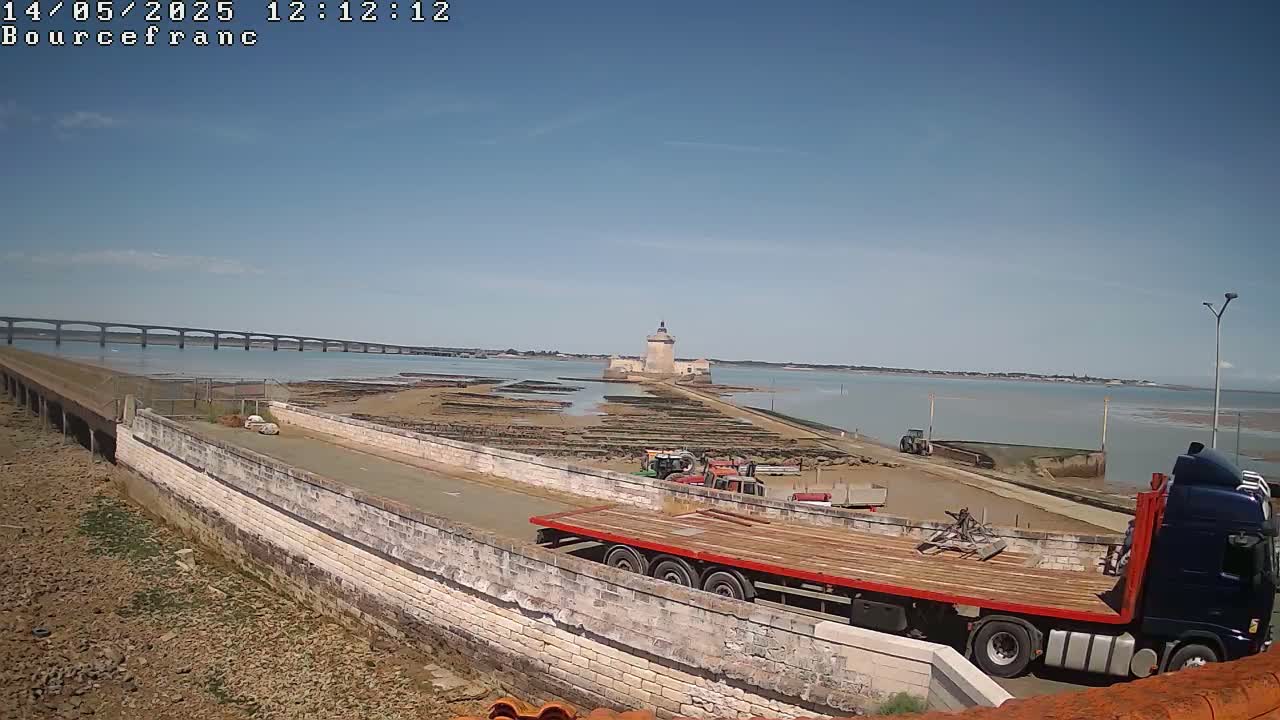 A flatbed truck is parked on a stone roadway overlooking a tidal flat with oyster beds, a distant stone tower, and a bridge under a clear blue sky.