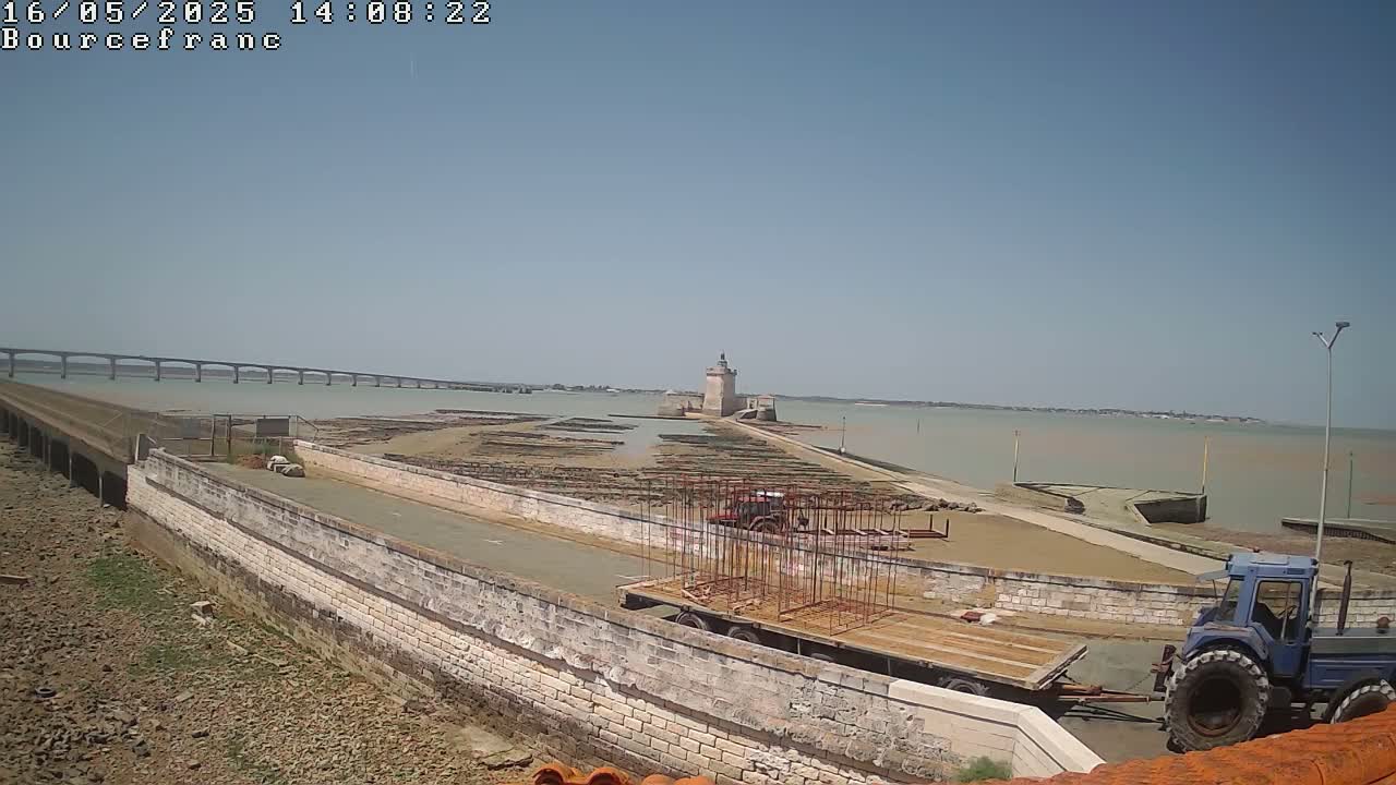 Under a clear blue sky, a stone causeway leads to a tower on a small island, with a long bridge visible in the distance and a tractor and trailer on the causeway.