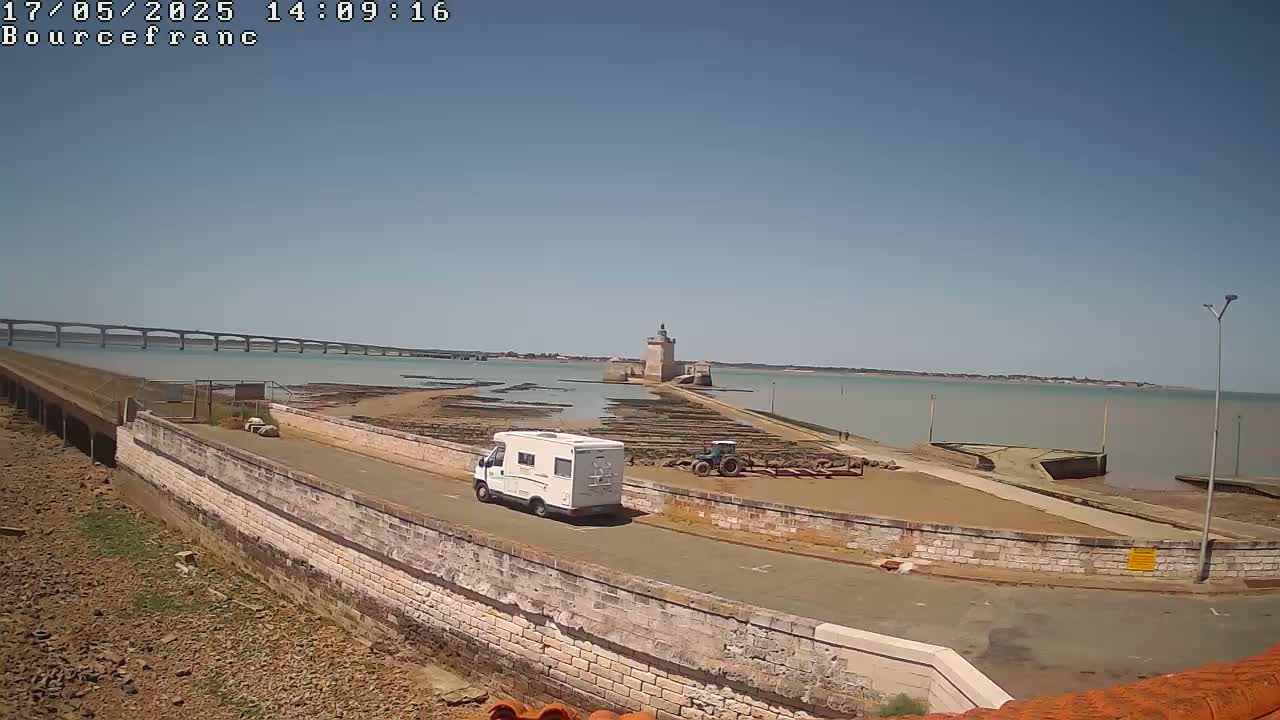 A white camper van drives along a road next to a stone wall overlooking a calm body of water, a stone tower, oyster beds, and a distant bridge under a clear blue sky.