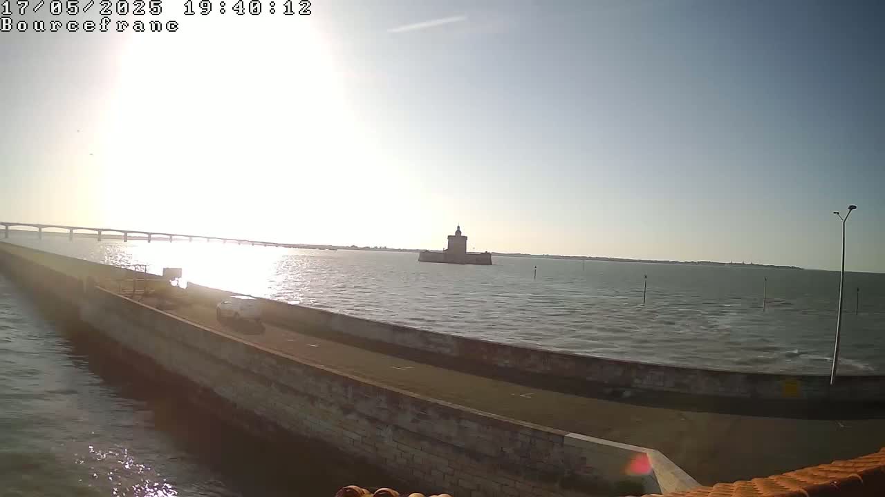 A long causeway extends into a calm body of water under a sunny sky, featuring a distant stone tower and a bridge in the background.