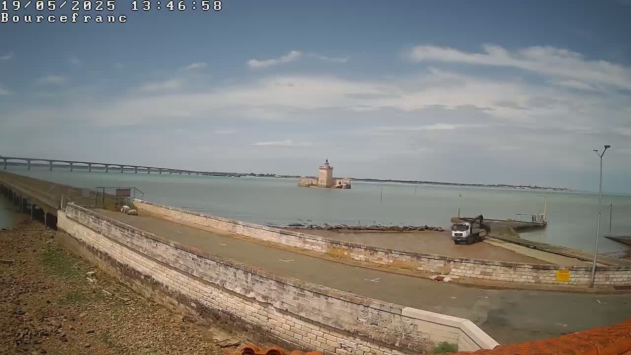 A partly cloudy day shows a stone structure and a truck near a body of water with a distant bridge under a partly cloudy sky.