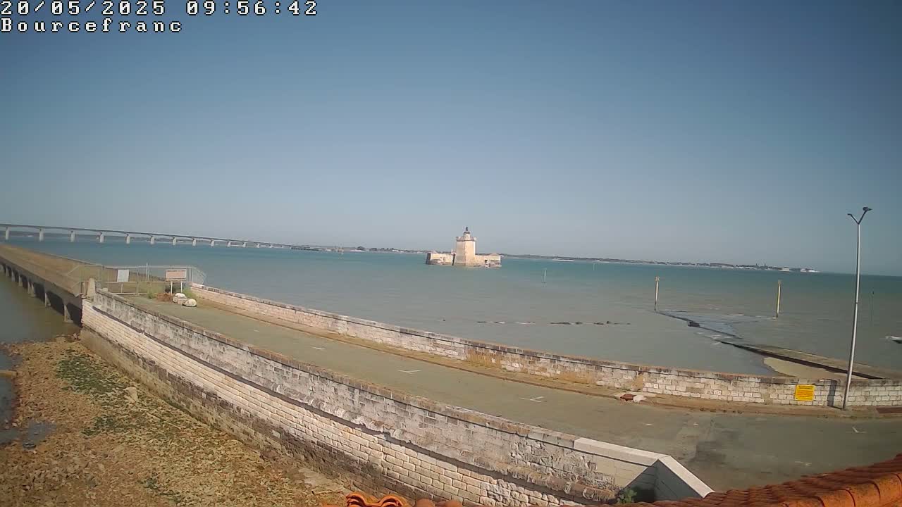 A stone roadway extends from a low wall into a calm body of water, with a distant bridge and a stone tower visible under a clear blue sky.