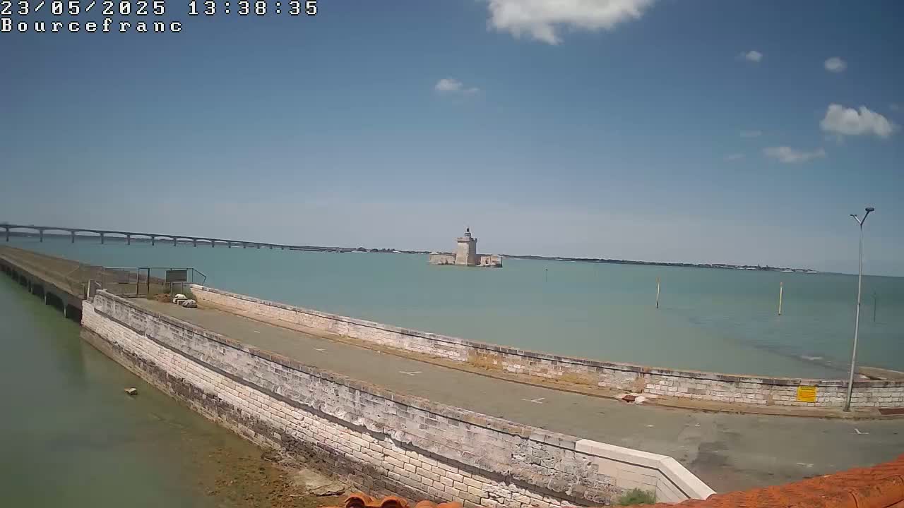 A stone wall with a road on top overlooks a calm, light-green body of water, a distant bridge, and a small stone tower in the middle of the water under a mostly sunny sky.