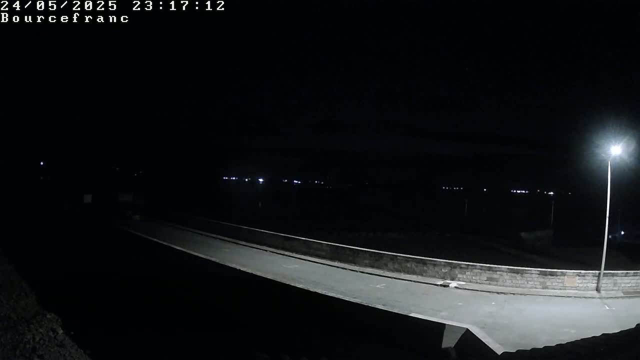 A nighttime, low-angle view shows a paved roadway curving across a low stone wall, extending into the distance where faint lights are visible.