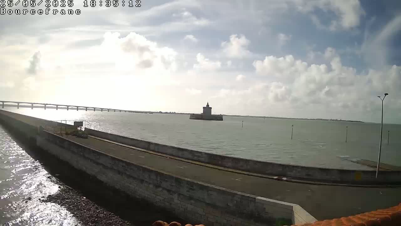 A long causeway extends into a body of water under a partly cloudy sky, with a distant tower visible in the middle of the water and a bridge in the far background.