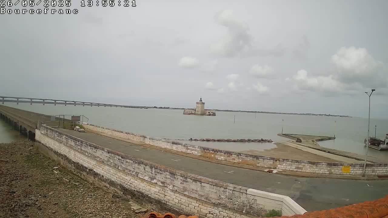 A stone structure and road curve around a body of water, under a cloudy sky, with a distant bridge visible.