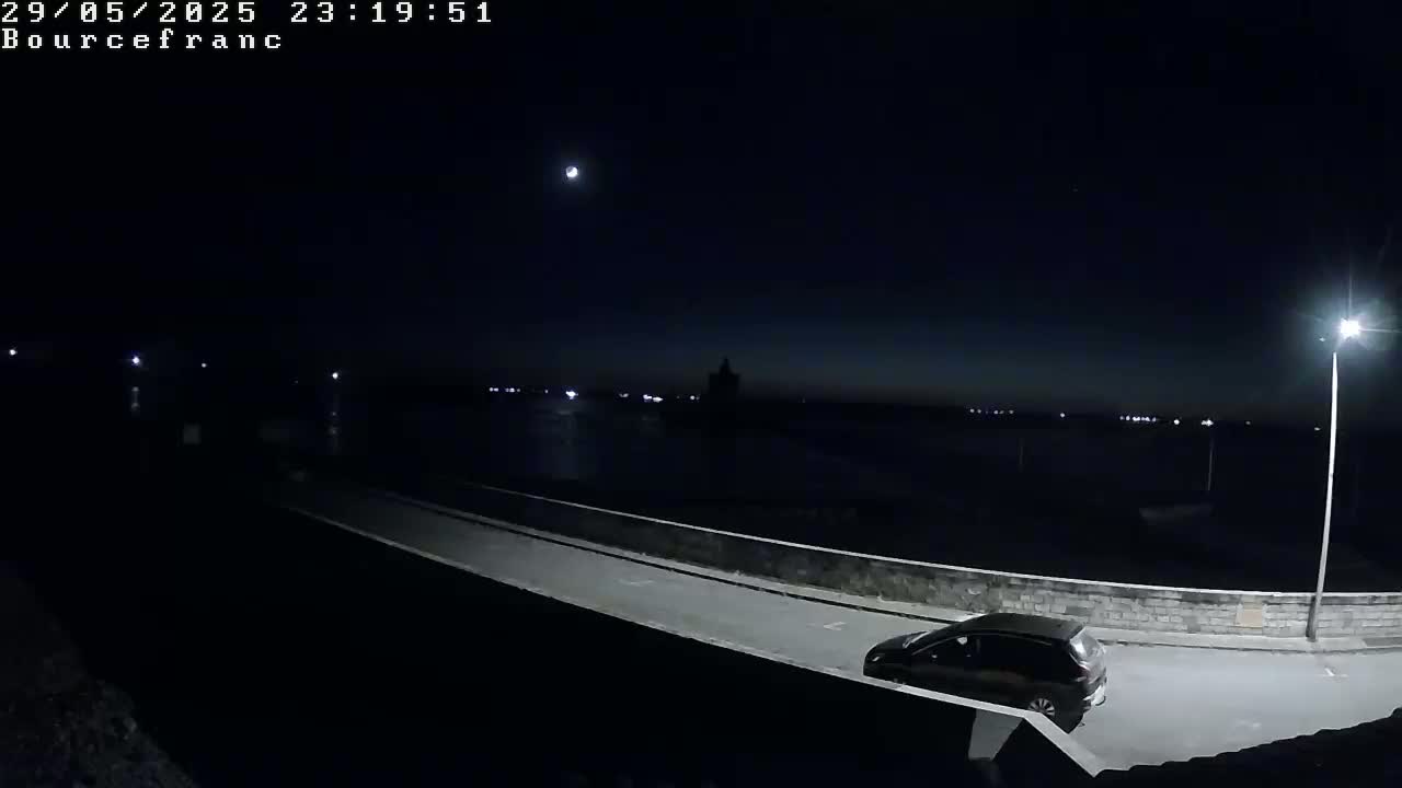 A dark-colored car is parked on a roadway next to a low wall overlooking a dark body of water under a mostly clear night sky.
