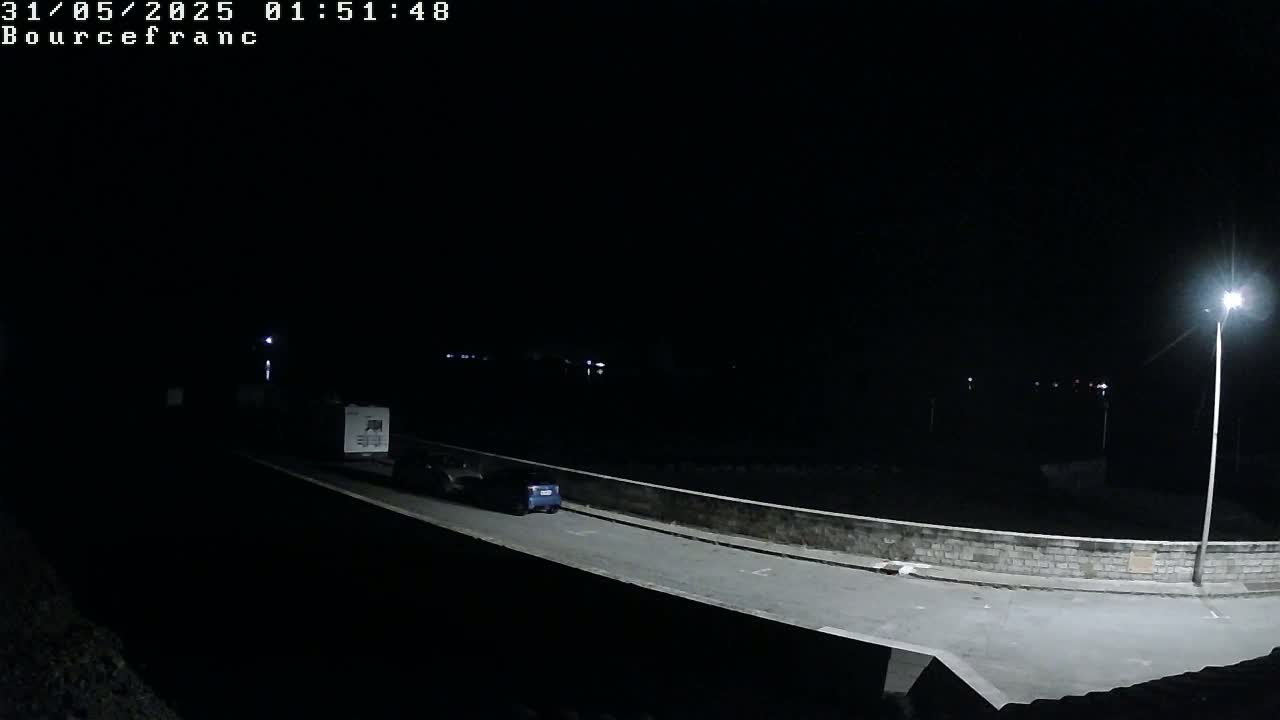 A dark, nighttime scene shows a blue car and a white van driving on a road lit by a streetlight, next to a low stone wall.