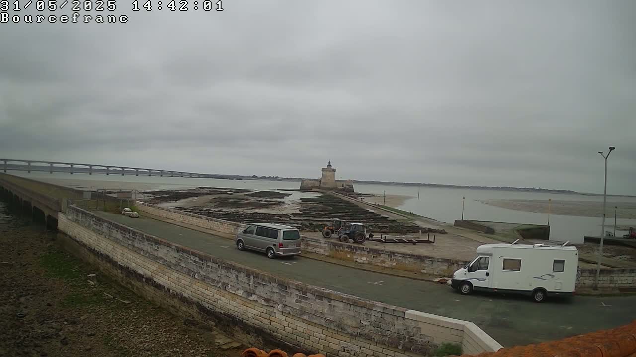 A gray and overcast day reveals a stone causeway leading to a fortified tower, with two vans and a tractor parked nearby, all set against the backdrop of a long bridge and a wide expanse of tidal flats.