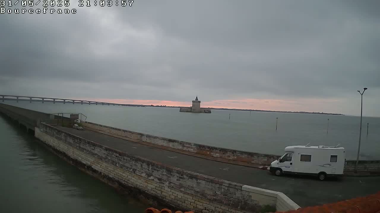 A white camper van is parked on a stone wall overlooking a calm body of water under an overcast sky, with a distant bridge and a stone tower visible.