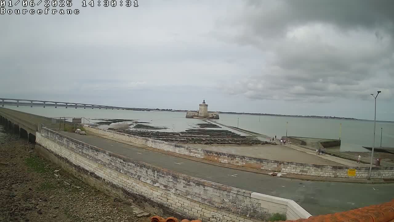 A stone tower sits on a tidal flat near a bridge and calm, cloudy water.