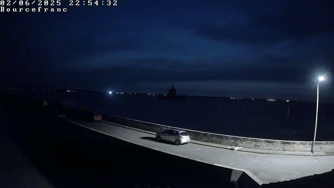 A silver car is parked on a dark road next to a low wall overlooking a calm body of water under a night sky with a few clouds.