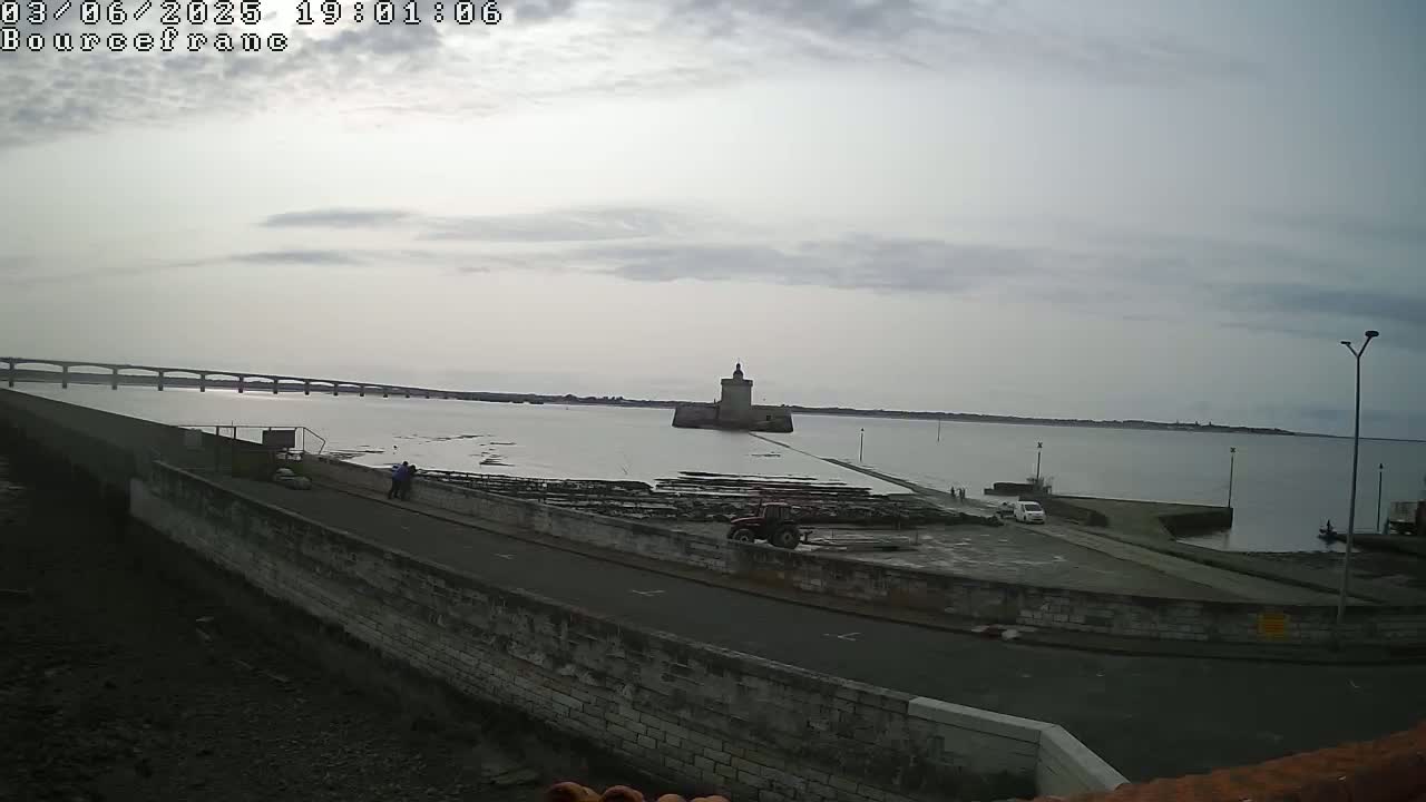 A stone tower sits in the middle of a calm, overcast bay, with a long bridge visible in the distance and a tractor parked nearby.