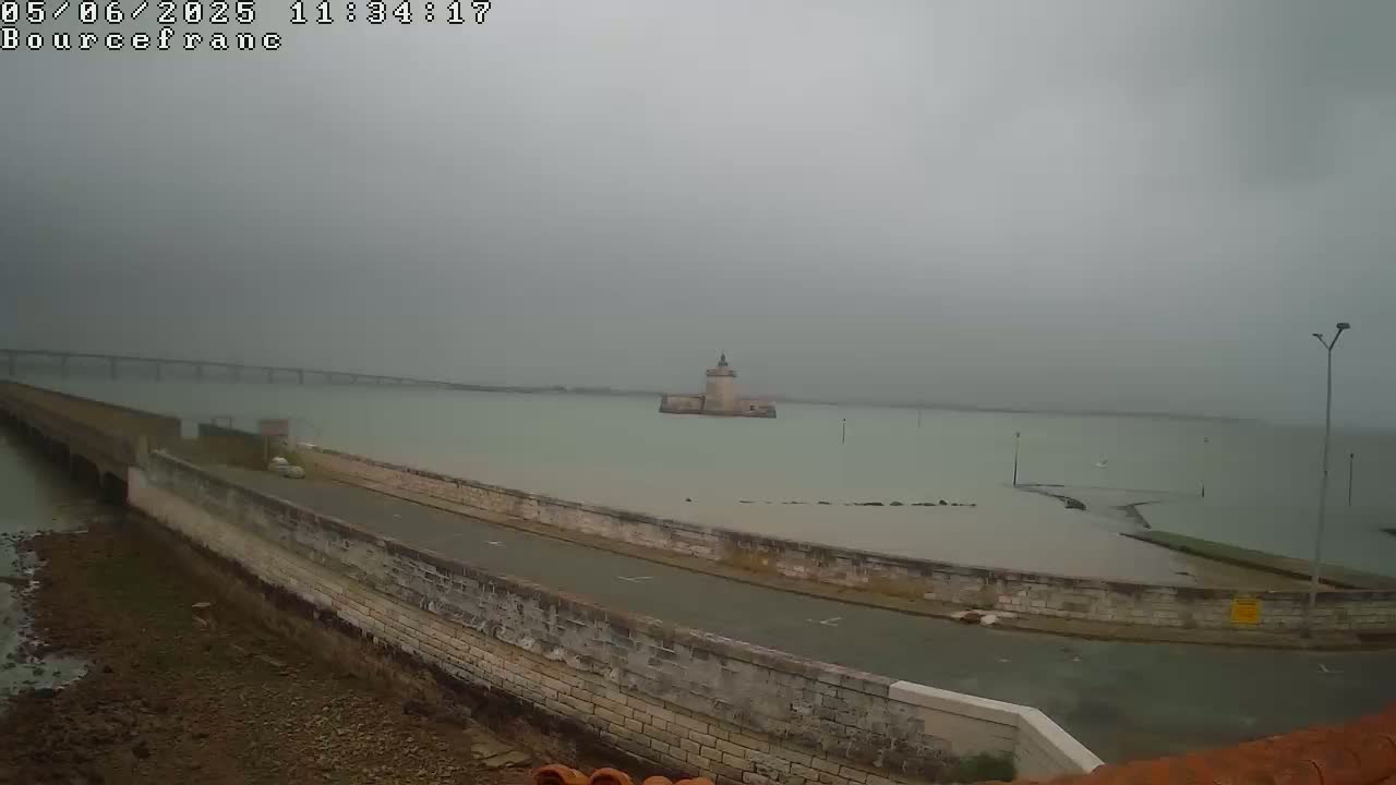 A long, low stone wall extends into a body of water on a foggy day, with a small tower visible in the distance and a bridge faintly in the background.