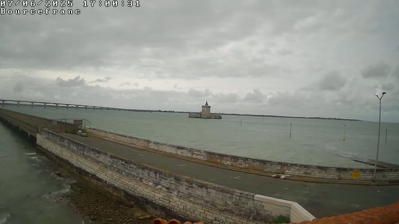 A stone wall curves along a roadway overlooking calm water, a distant bridge, and a small, stone tower in the overcast sky.