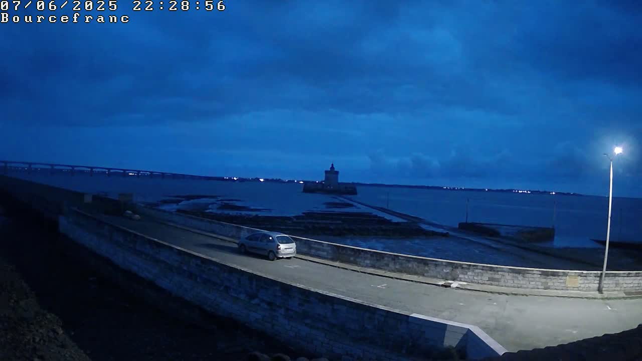 A silver car drives along a coastal road at twilight, passing a stone wall with a distant tower and bridge visible across the water under a dark blue, cloudy sky.