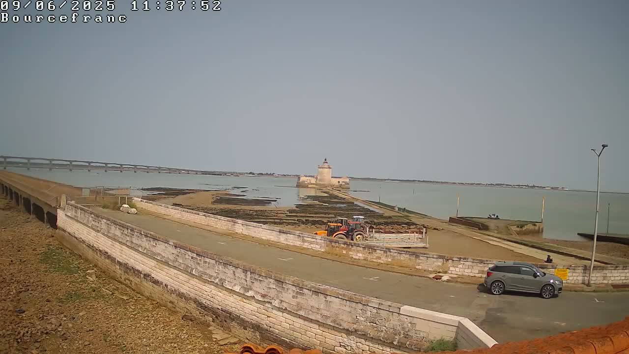 A gray SUV is parked near a stone wall overlooking a causeway leading to a stone tower on a calm, sunny day, with a bridge visible in the distance.