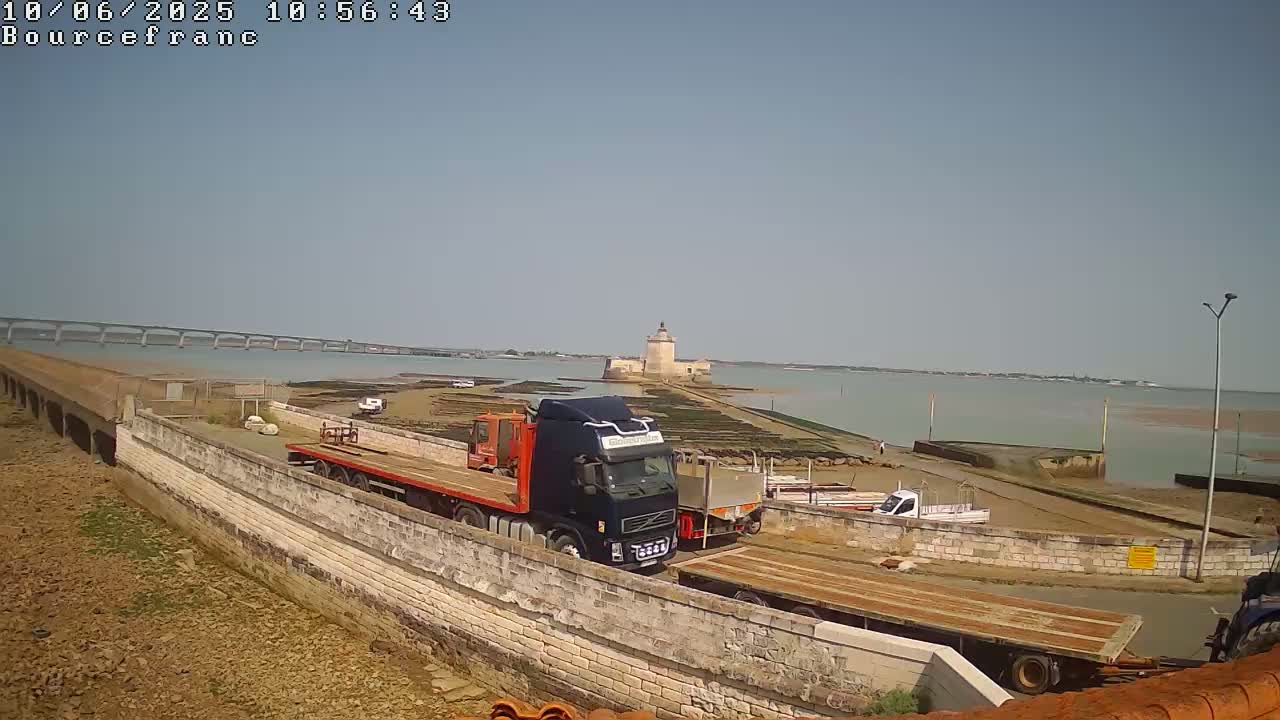 A sunny day reveals several trucks parked near a stone wall overlooking a body of water, a distant bridge, and a stone tower on an island.