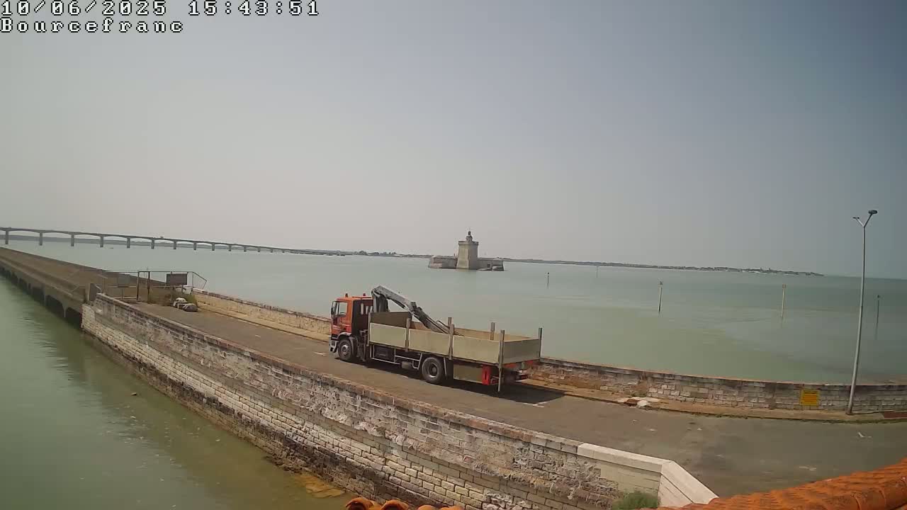 An orange truck with a crane and open-top container drives along a stone seawall, with a long bridge and a stone tower visible in the distance across calm, light green water under a hazy sky.