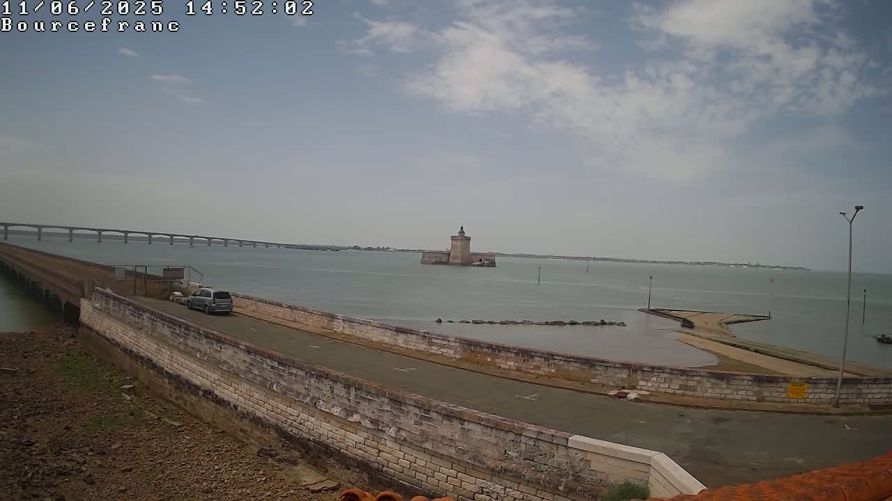 A long stone causeway leads to a small island with a stone tower, a bridge spans the distance in the background, under a partly cloudy sky.
