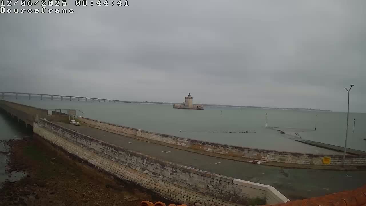A stone roadway curves around a body of water under an overcast sky, with a distant bridge and a small tower visible in the background.