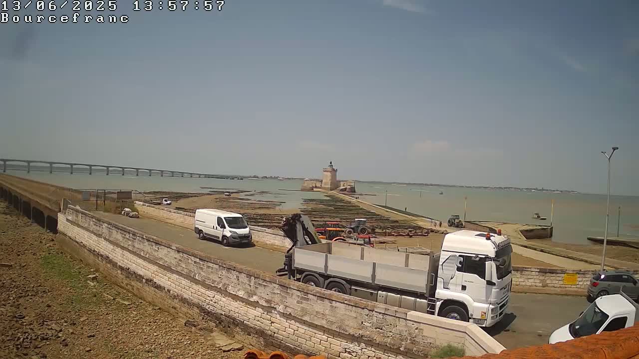 Under a clear, sunny sky, a coastal scene shows a stone wall, several trucks, a van, and a stone tower on an island connected to the mainland by a causeway, with a long bridge visible in the distance.