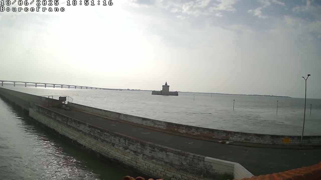 A stone seawall curves around a body of water, with a distant bridge and a small, fortified island visible under a partly cloudy sky.