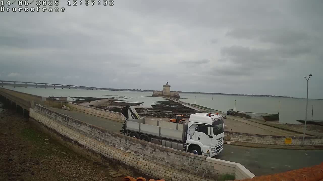 A white truck with a crane is parked near a stone wall overlooking a body of water on an overcast day, with a distant bridge and a stone tower visible in the background.