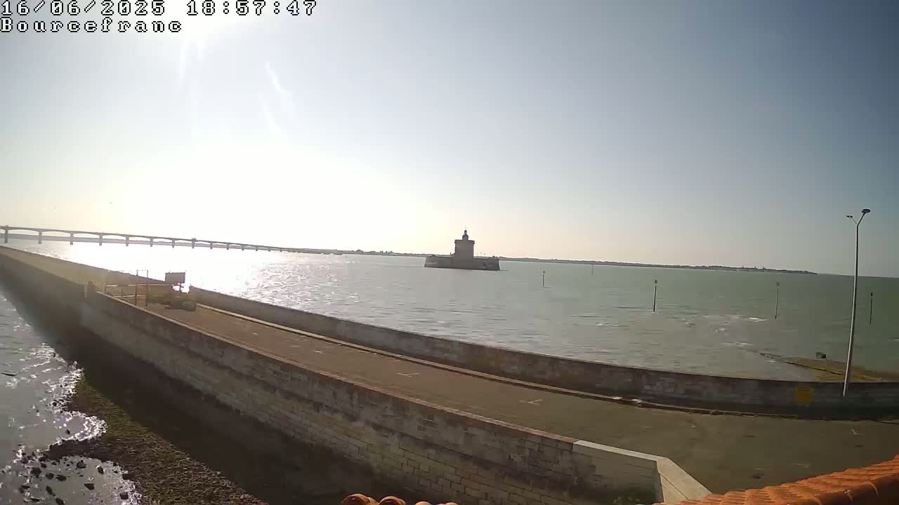 A stone wall extends into a calm body of water, with a distant bridge and a small stone tower visible under a bright, sunny sky.