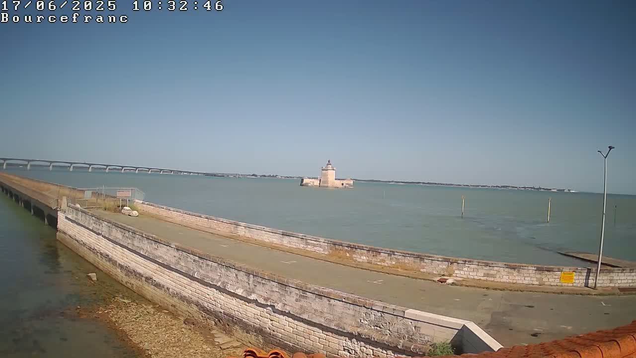 A stone wall extends into calm, light-green water towards a small, stone tower, with a long bridge visible in the distance under a clear blue sky.