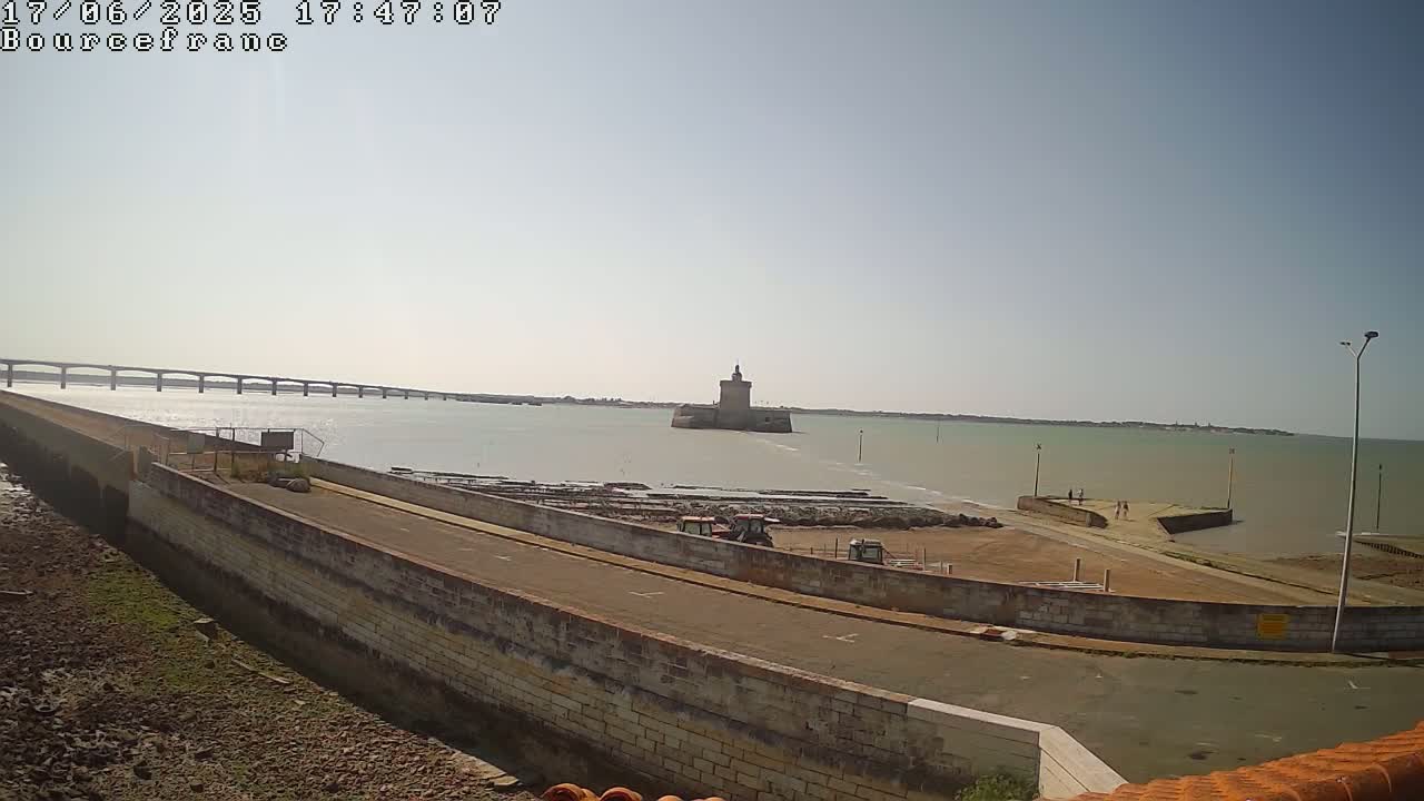 A stone wall and paved area overlook a calm body of water with a distant bridge and a stone tower on an islet under a clear, sunny sky.
