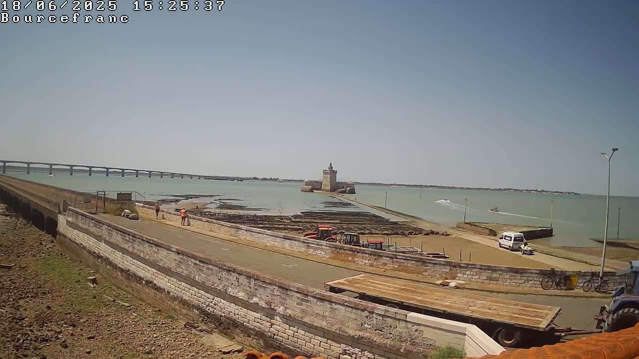 A coastal scene on a sunny day shows a stone structure with a tower in the water, a long bridge in the distance, and construction equipment near a roadway.