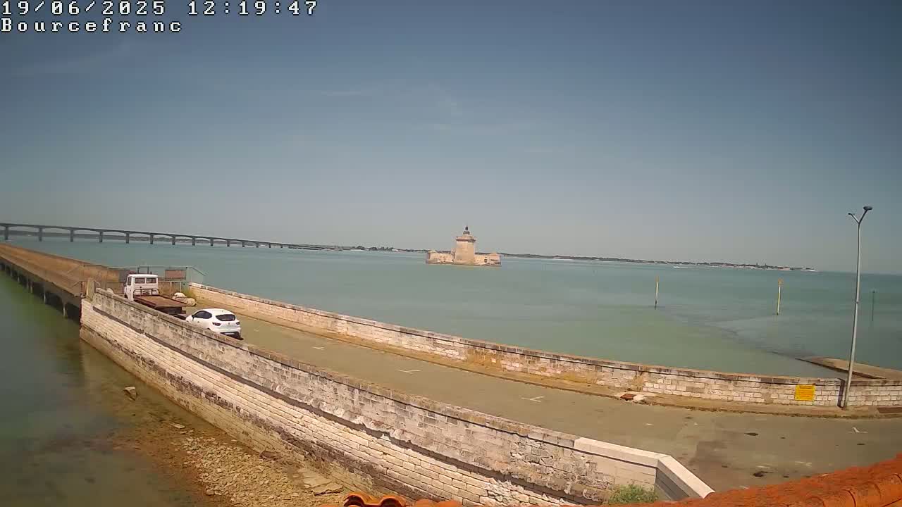 A long causeway extends into a calm, light-green body of water toward a stone tower; a distant bridge is visible in the background under a clear, sunny sky.