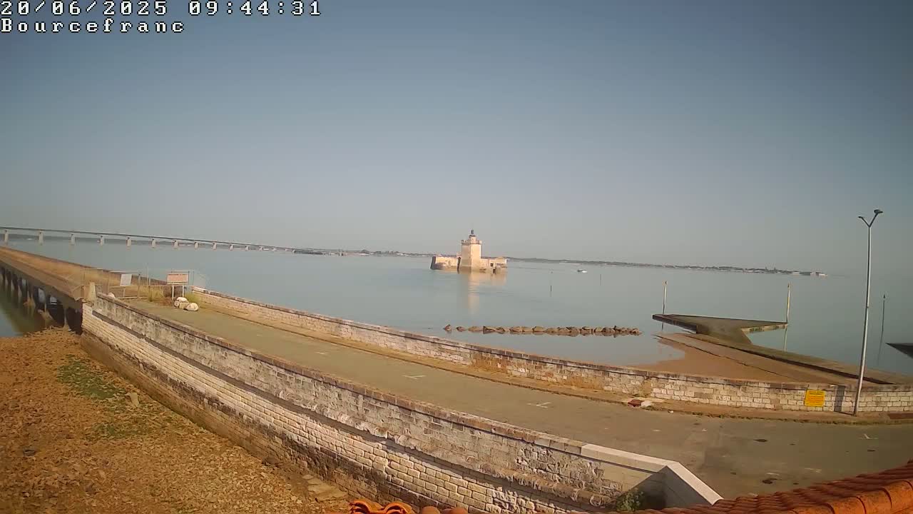 A stone causeway extends into calm, light-blue water toward a small stone tower, with a long bridge visible in the distance under a clear, bright sky.