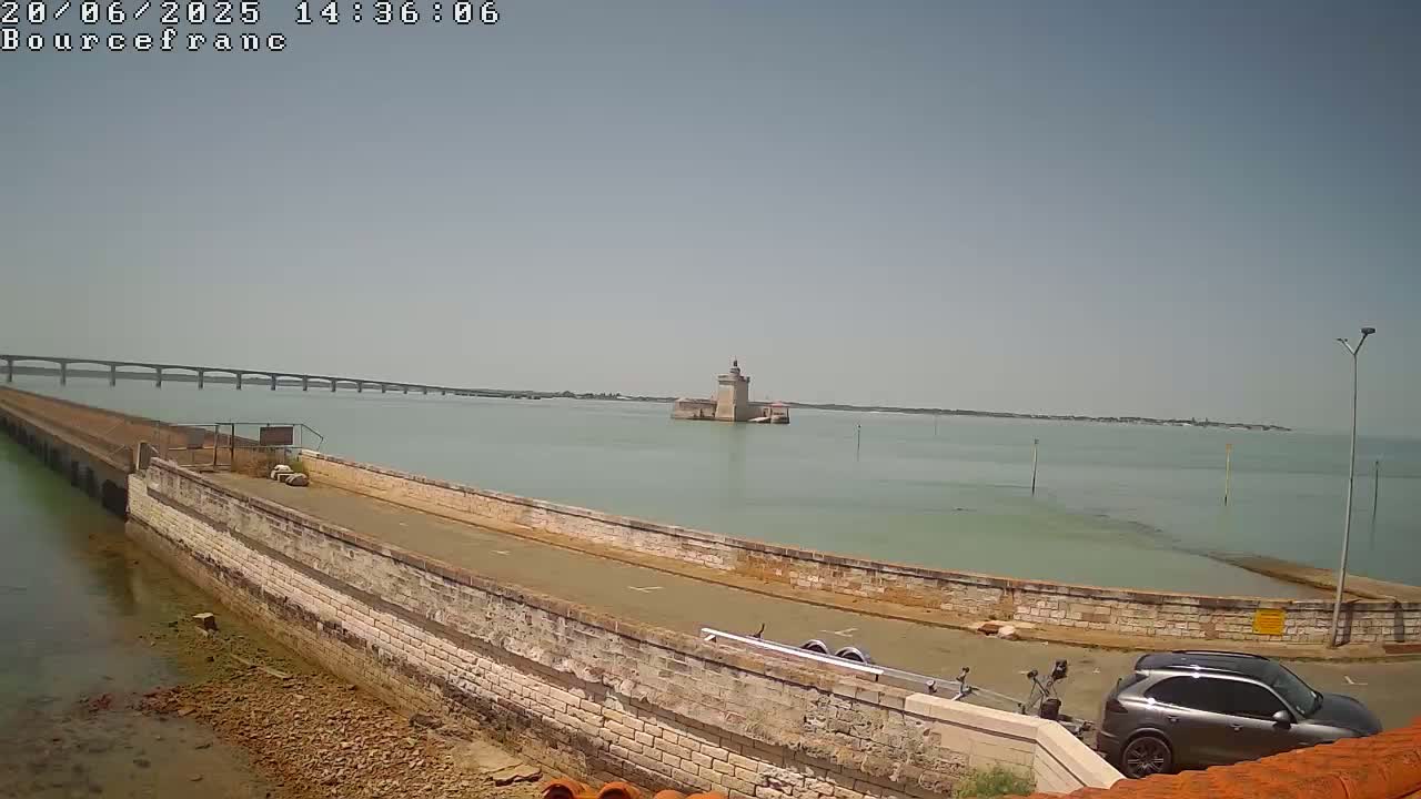 A stone roadway extends toward a distant island fortress on calm, light-green water under a clear, bright sky, with a long bridge visible in the background.
