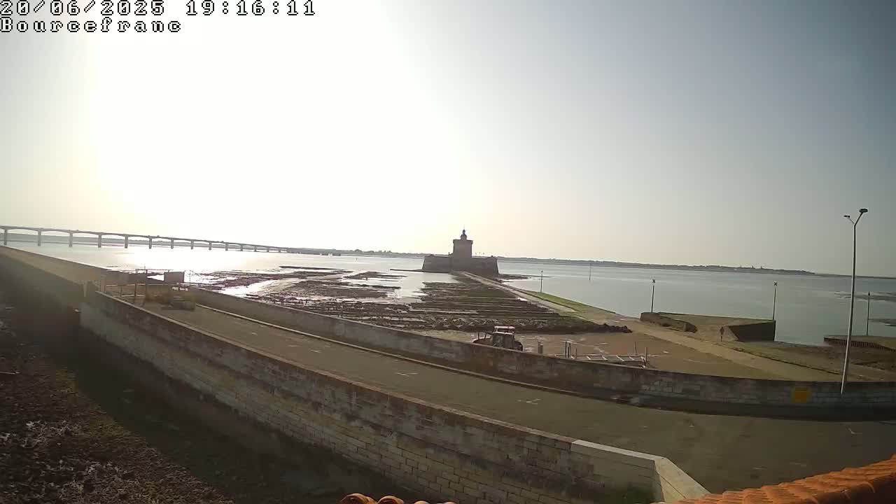 A stone tower sits on a tidal flat near a long bridge and calm body of water under a bright, hazy sky.