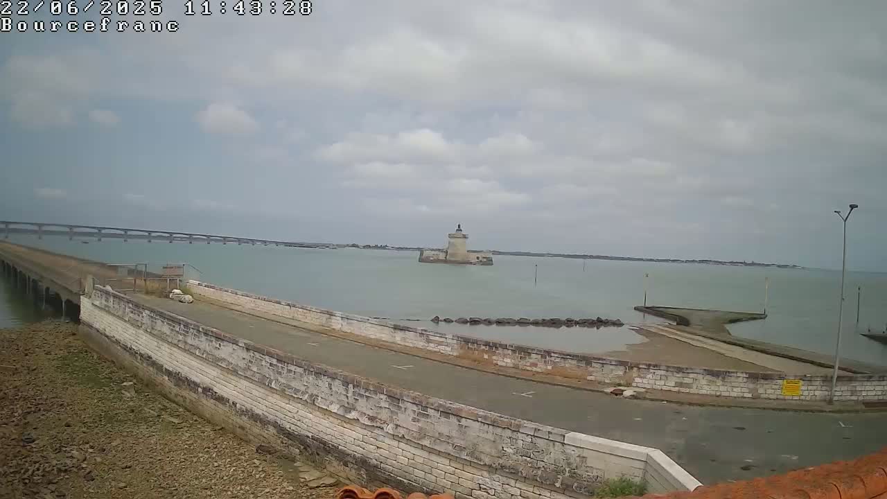 A stone seawall curves around a small paved area next to calm, light-green water, with a distant bridge and a small stone tower visible under a mostly cloudy sky.