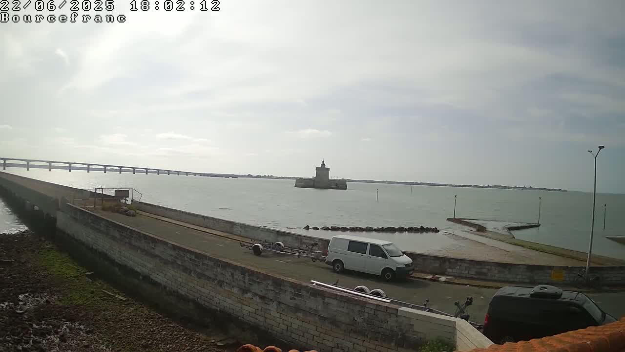 A white van with a boat trailer is parked on a stone roadway next to a body of water, with a distant bridge and a stone tower visible under a cloudy sky.