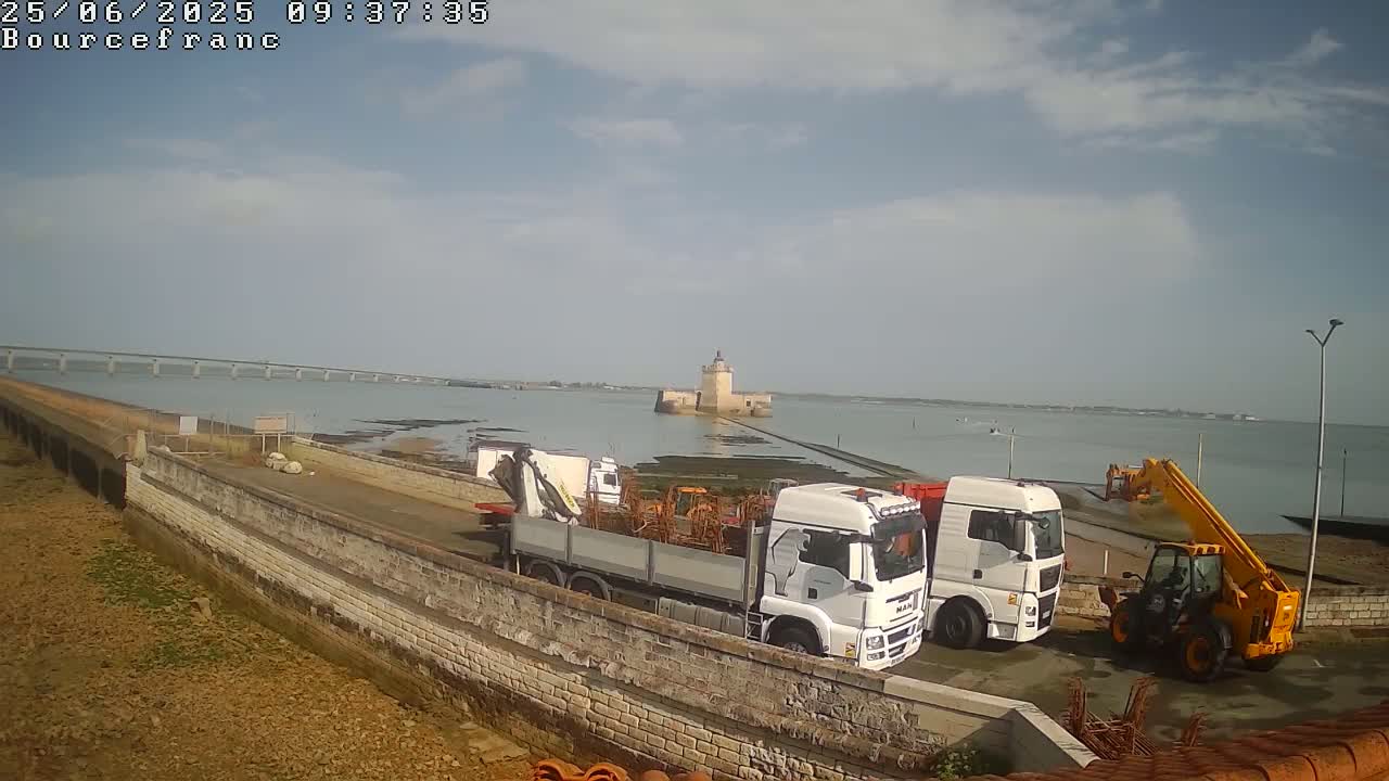 Under a partly cloudy sky, two large white trucks, a yellow telehandler, and a stone wall are situated near a body of water with a distant bridge and a stone tower.