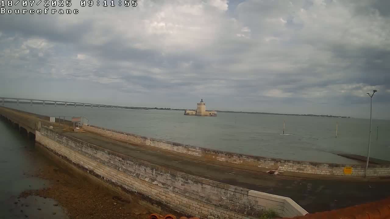 A stone seawall curves alongside a calm, gray body of water, featuring a distant tower and a bridge under a cloudy sky.