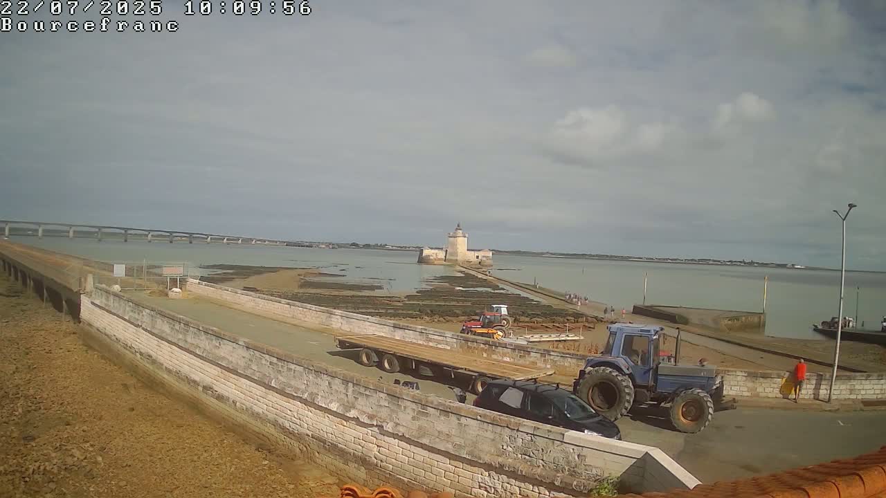 A stone causeway leads to a small island with a stone tower, under a partly cloudy sky, with construction vehicles and people present.