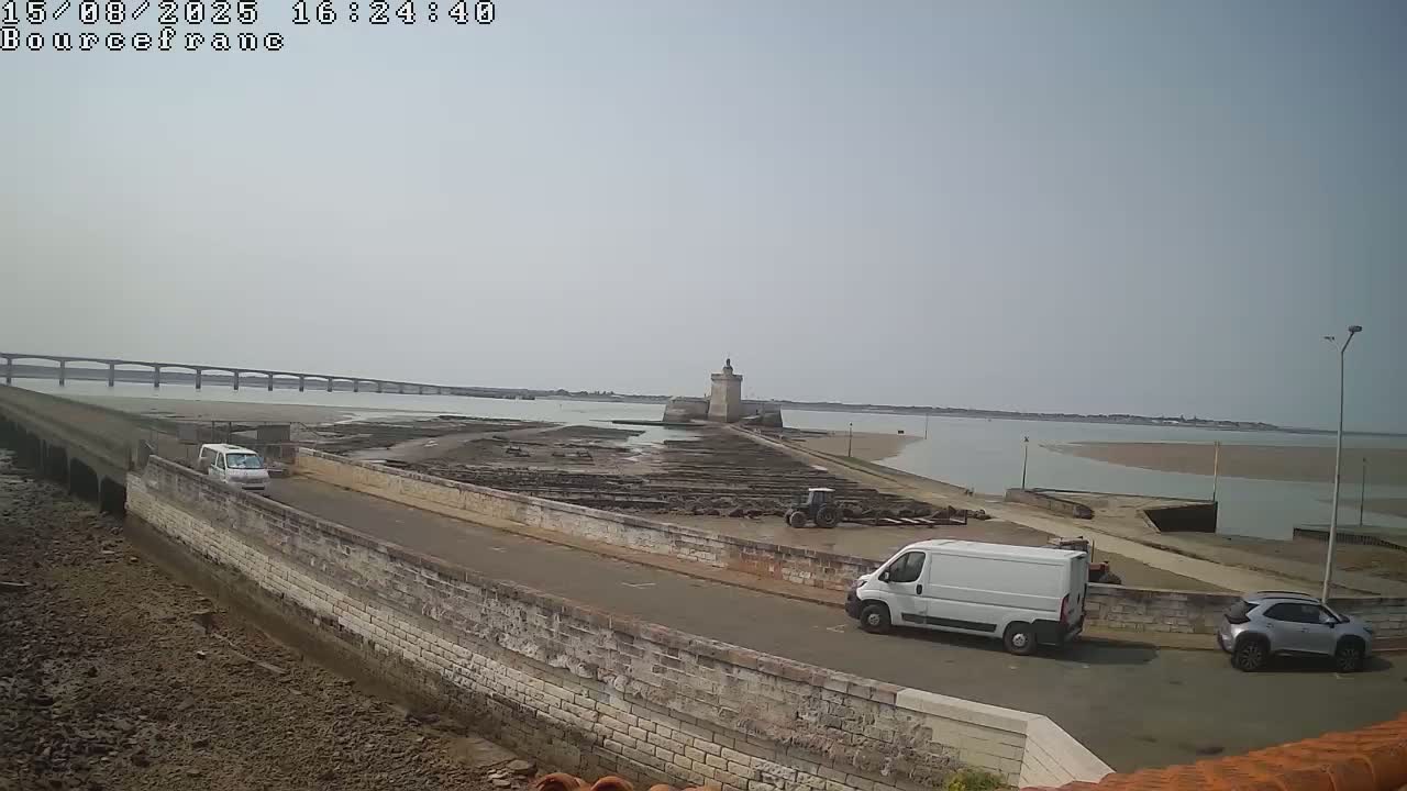 A stone causeway leads to a small island with a stone tower, under a partly cloudy sky, with construction vehicles and people present.