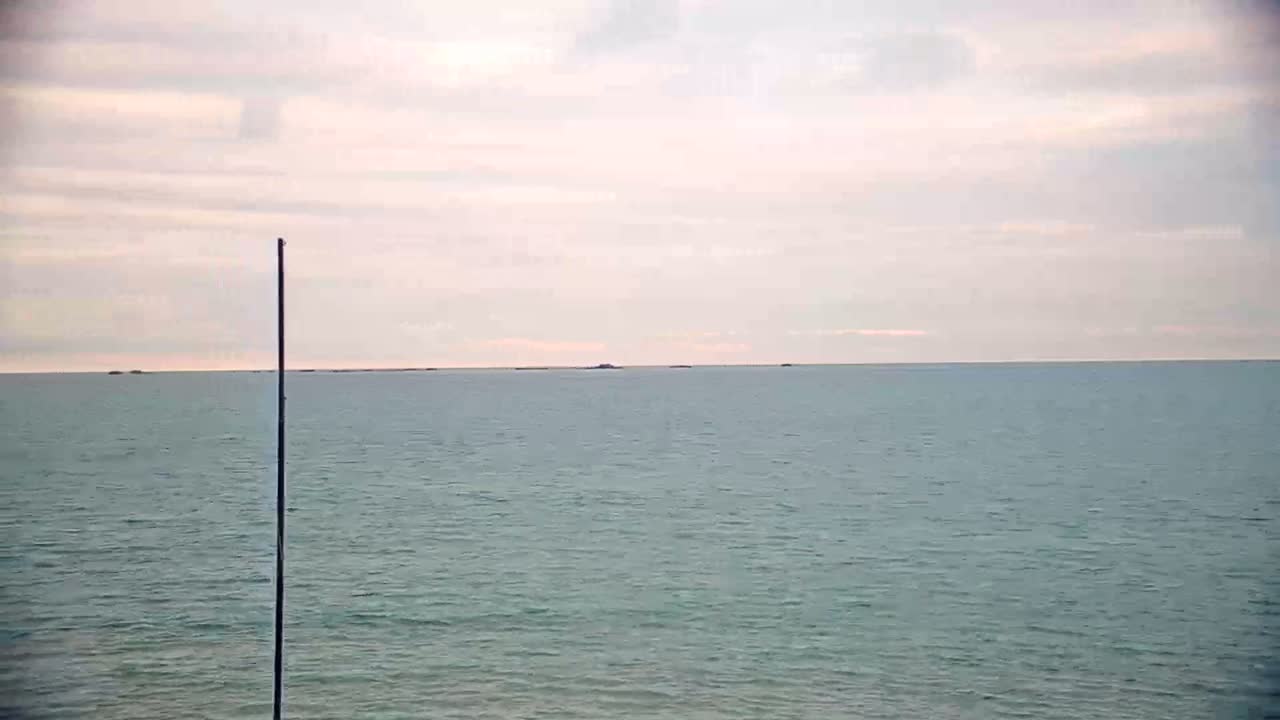 A calm, pale-sky day is shown over a tranquil body of water, with a tall, thin pole in the foreground.