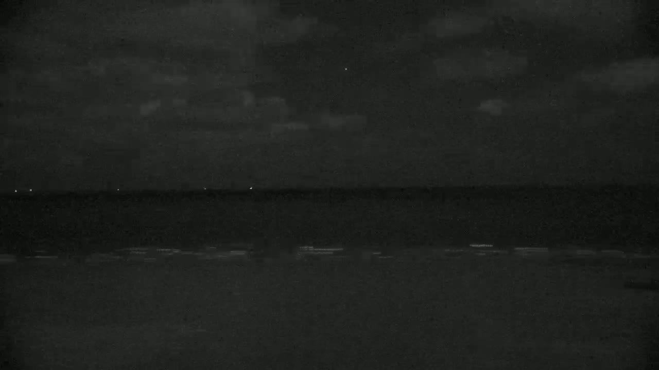 A dark, nighttime scene shows a calm body of water under a cloudy sky, with a few faint lights visible on the horizon.
