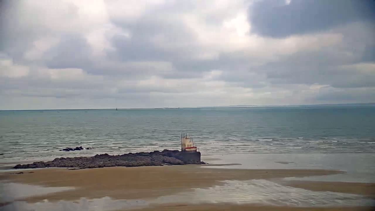 A sandy beach stretches to a calm sea under a cloudy sky, with a small structure perched on rocks near the shore.