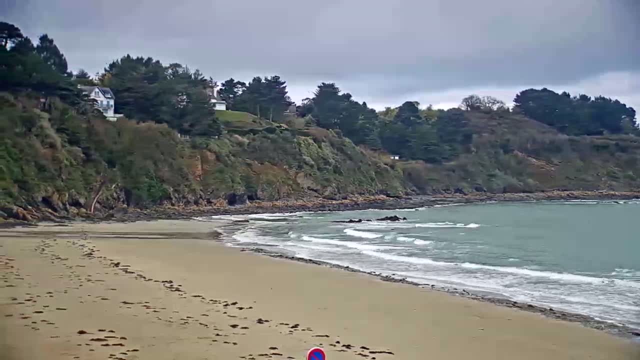 A sandy beach with small waves laps against the shore, under a cloudy sky,  backed by a rocky cliff covered with green vegetation and houses.